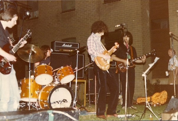 A five-piece band playing outdoors at night in front of a brick building, lit by stage lights, with a Marshall stack and drum kit.