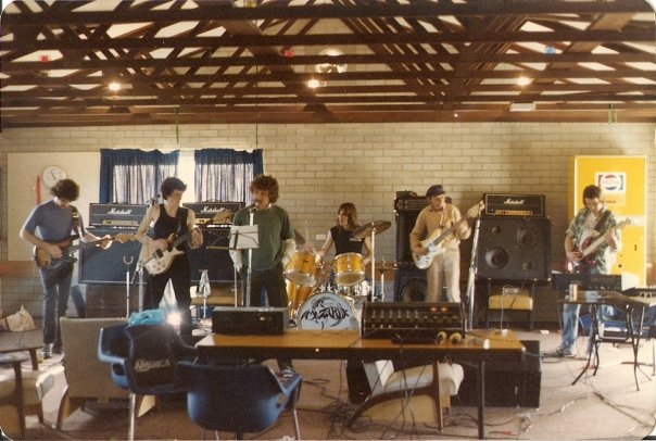 Six musicians rehearsing in a hall with exposed roof beams, surrounded by amplifiers, a drum kit and stacked chairs.