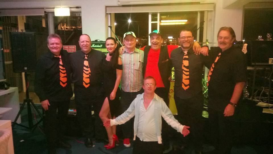 Eight performers posed together after a gig, several wearing black shirts with orange tiger-stripe ties, one crouching at the front with arms outstretched.